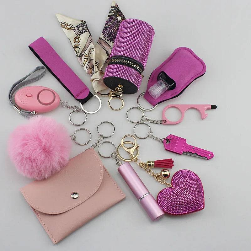 Complete set of SafetyKey accessories including a personal alarm with siren, cardholder, wristband, lipstick holder, perfume bottles, organizer pouch, fluffy pompom, whistle, and other keychain items in pink and purple tones.