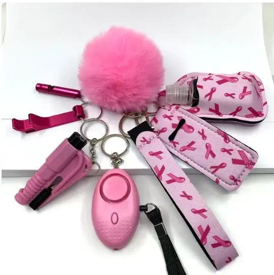 Eight-piece SafetyKey set, including a personal alarm with LED light, bottle protective sleeve, door opener artifact, two lipstick holders, pom-pom ball, sanitizer holder, keychain, and a wrist strap, all in pink with ribbon designs.